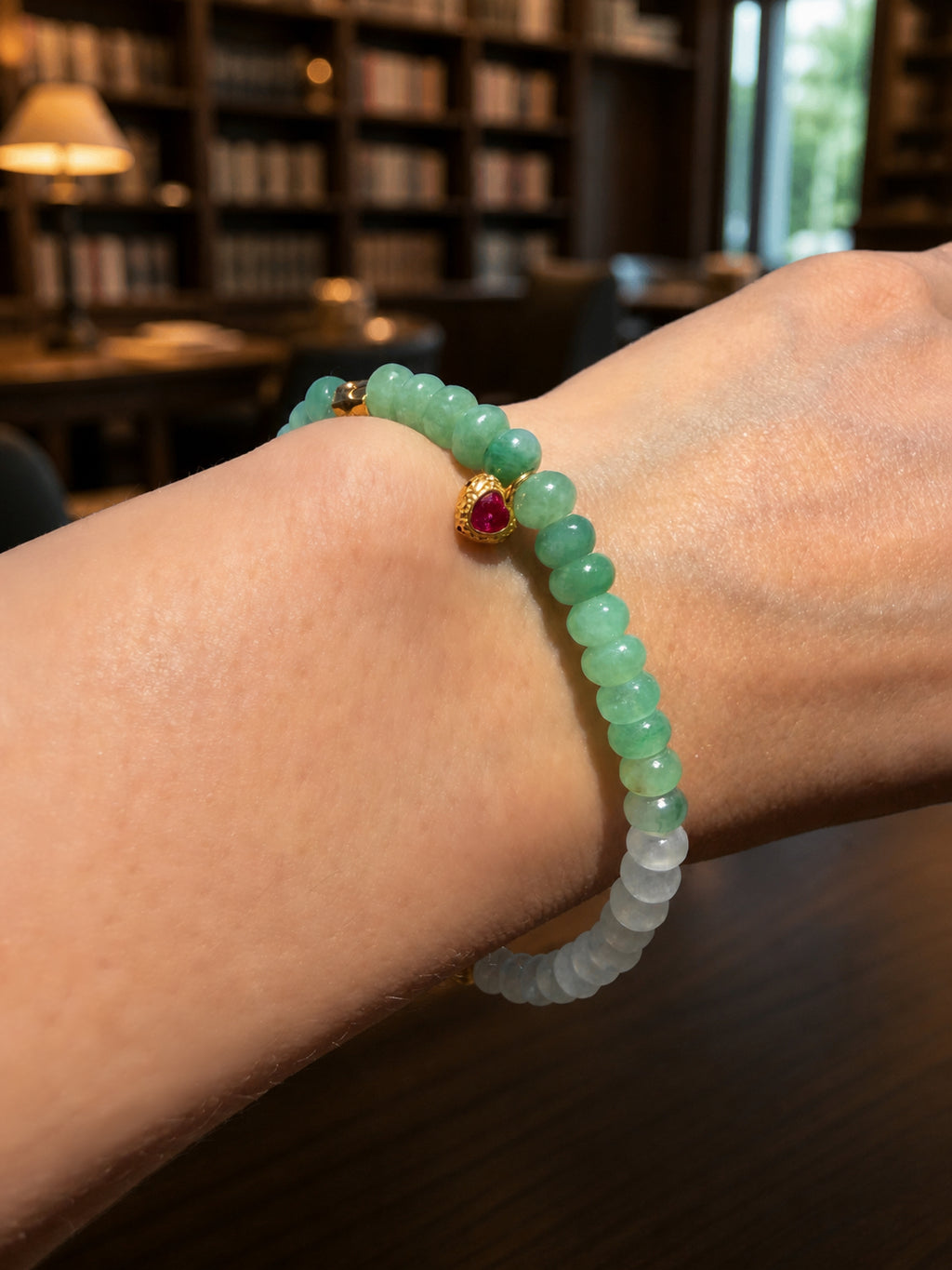 Jadeite beaded bracelet on wrist with green beads, gold clasp, and small red gemstone charm