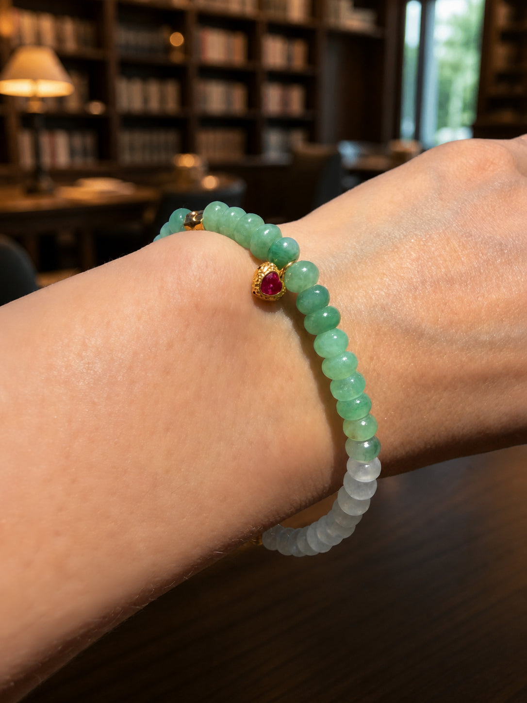Jadeite beaded bracelet on wrist with green beads, gold clasp, and small red gemstone charm
