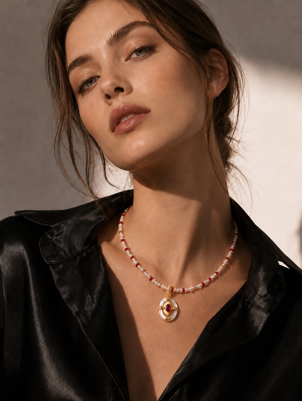Beaded gold necklace with oval red-and-pearl pendant, worn on a black shirt